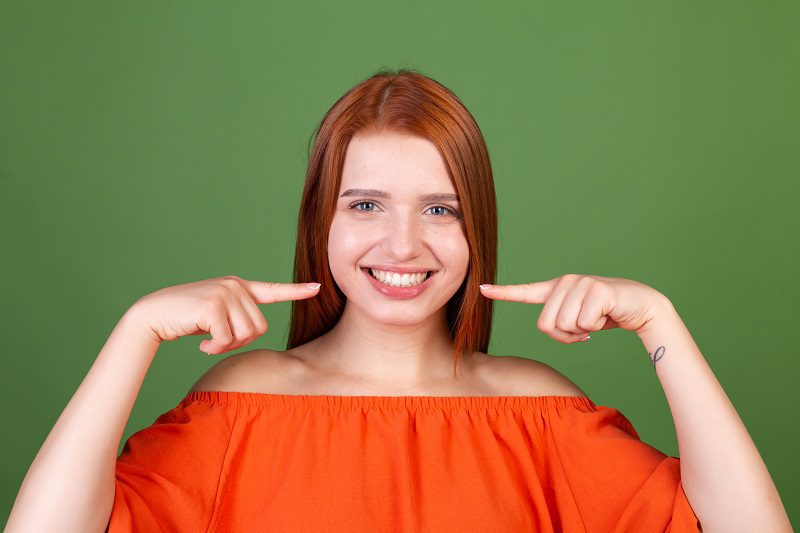 young-red-hair-woman-casual-orange-blouse-green-wall-point-fingers-white-teeth-perfect-smile.jpg